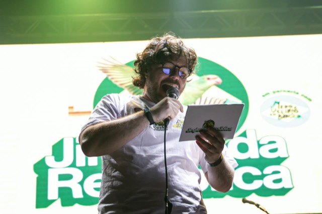 Apresentador de camiseta branca lê um cartão ao microfone, diante do telão com o logo do campeonato.
