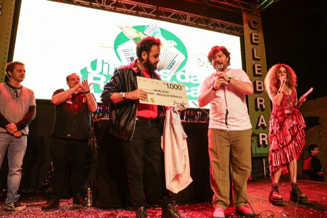 No palco, um premiado de jaqueta de couro segura o cheque de R$ 1.000 ao lado dos apresentadores.