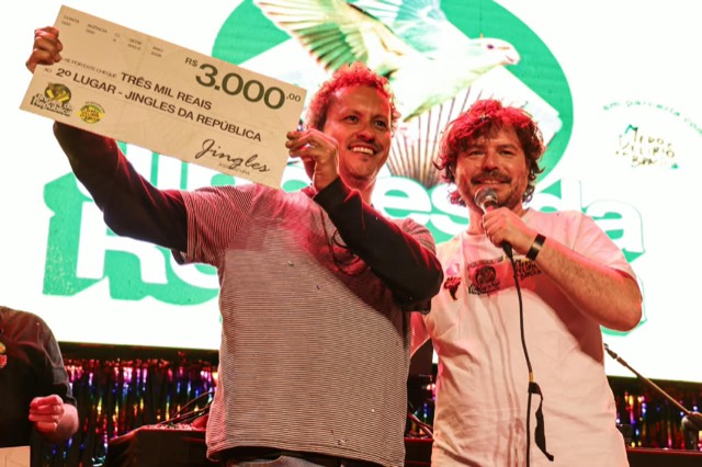 Premiado mostra o cheque de R$ 3.000 enquanto o apresentador fala ao microfone.