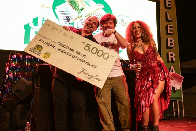 Premiados erguem o cheque de R$ 5.000 do 1º lugar, ao lado da apresentadora.