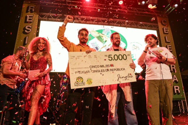 Premiado ergue o punho ao lado do cheque de R$ 5.000 do 1º lugar, sob confete.